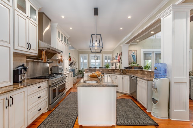 kitchen with stainless steel appliances, a sink, ornamental molding, wall chimney range hood, and dark stone counters