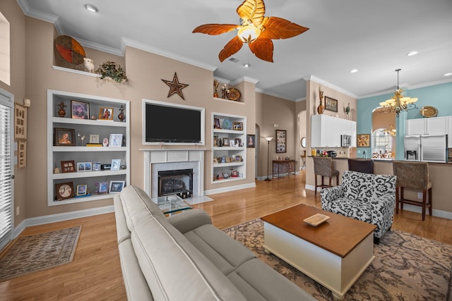 living room with ceiling fan with notable chandelier, built in shelves, light wood-style floors, and a high end fireplace