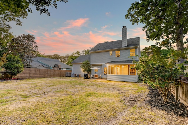 back of property at dusk with a yard, a chimney, a fenced backyard, and a sunroom