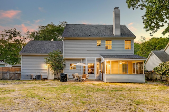 back of property at dusk with a shingled roof, a sunroom, a fenced backyard, a chimney, and a lawn