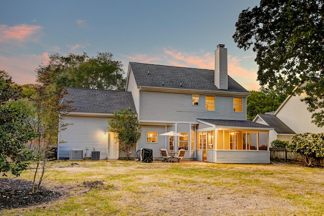back of property at dusk featuring a chimney, a lawn, a sunroom, central air condition unit, and fence