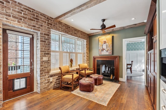 sitting room with a multi sided fireplace, beamed ceiling, ceiling fan, brick wall, and hardwood / wood-style floors