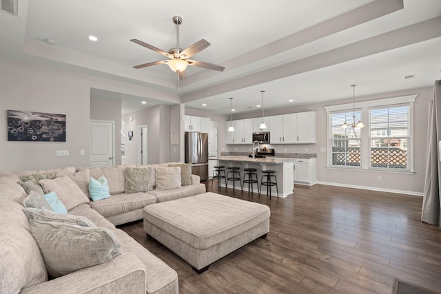 living room with sink, ceiling fan with notable chandelier, dark wood-type flooring, and a tray ceiling