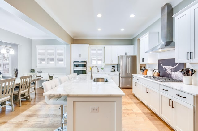 kitchen featuring light wood finished floors, appliances with stainless steel finishes, a sink, wall chimney range hood, and a kitchen breakfast bar