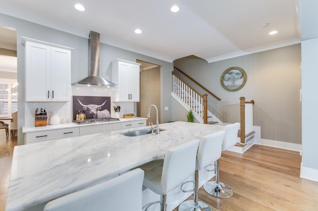 kitchen with light stone counters, a sink, wall chimney range hood, light wood-type flooring, and stainless steel gas stovetop