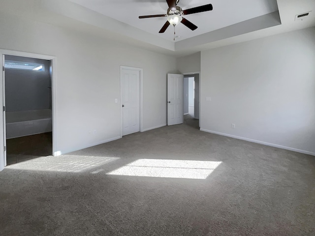 unfurnished bedroom with ceiling fan, ensuite bath, a raised ceiling, and dark colored carpet