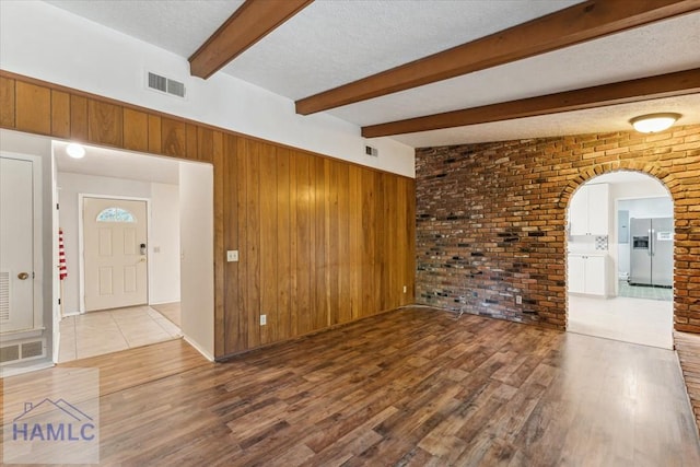 unfurnished living room featuring brick wall, a textured ceiling, wood-type flooring, beam ceiling, and wood walls