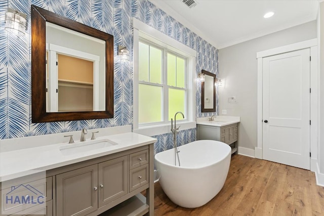 bathroom with wood finished floors, wallpapered walls, two vanities, a freestanding tub, and a sink