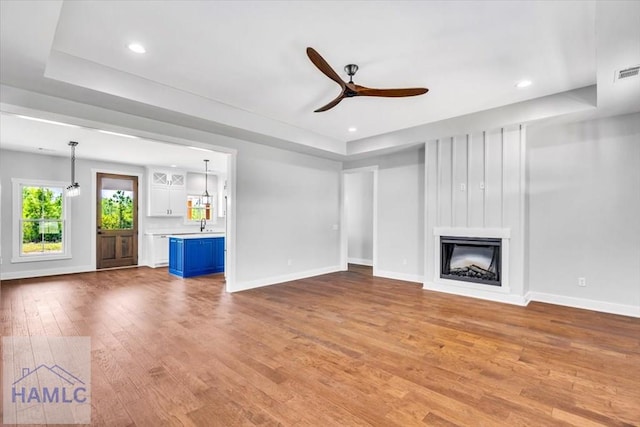 unfurnished living room featuring wood-type flooring, a ceiling fan, baseboards, a fireplace, and a tray ceiling