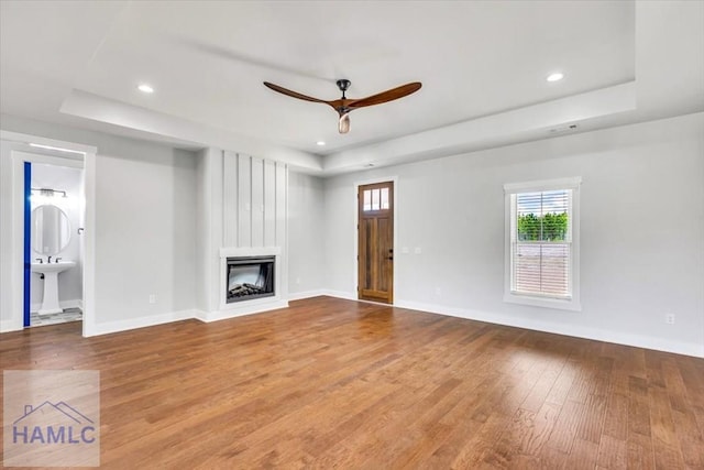 unfurnished living room with a tray ceiling, a large fireplace, wood finished floors, ceiling fan, and baseboards
