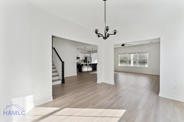 unfurnished living room featuring light wood-type flooring, stairway, baseboards, and ceiling fan with notable chandelier