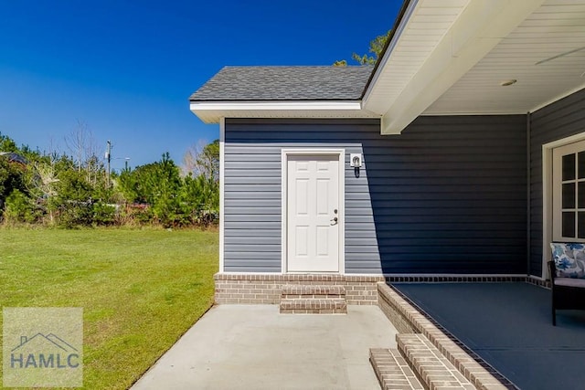 property entrance featuring a shingled roof, a patio, a lawn, and brick siding