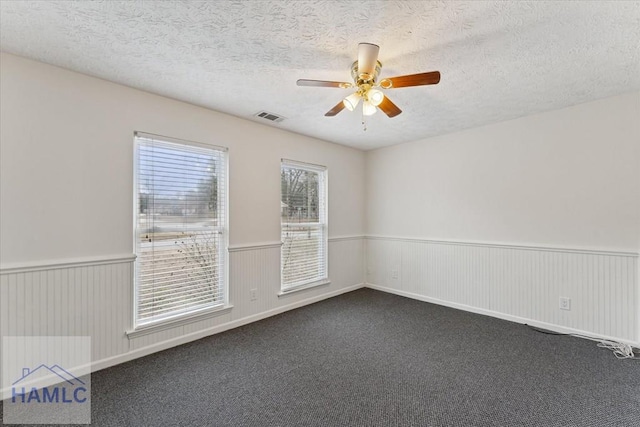 unfurnished room with a textured ceiling, ceiling fan, visible vents, wainscoting, and dark colored carpet