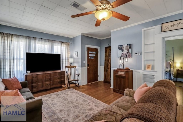 living area with built in shelves, wood finished floors, a wainscoted wall, visible vents, and ornamental molding