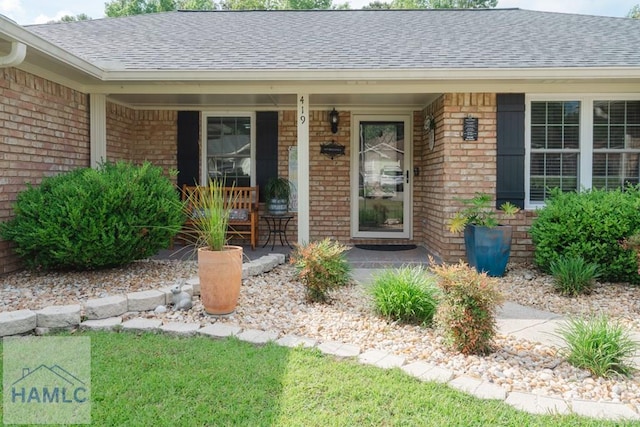 view of exterior entry with a shingled roof, covered porch, and brick siding