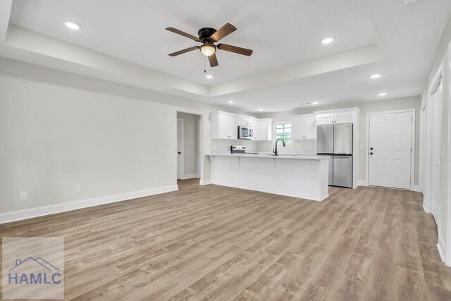 unfurnished living room with ceiling fan, light wood-type flooring, sink, and a tray ceiling