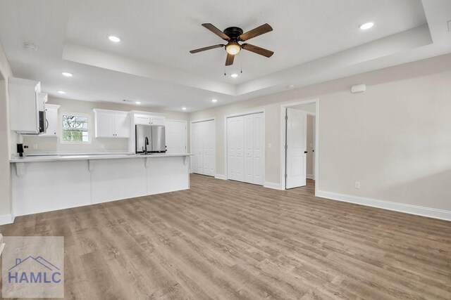 unfurnished living room with ceiling fan, a raised ceiling, sink, and light hardwood / wood-style flooring