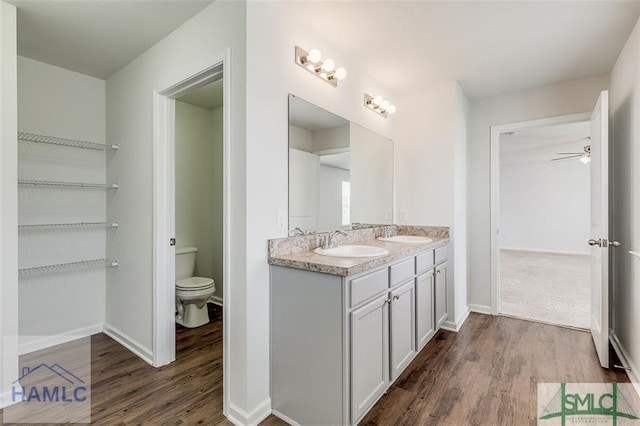 bathroom with wood finished floors, toilet, double vanity, a ceiling fan, and baseboards