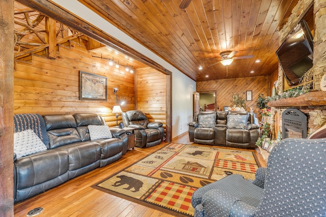 living room featuring wood walls, wooden ceiling, ceiling fan, a stone fireplace, and light wood-type flooring