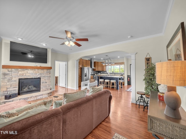 living room featuring ceiling fan with notable chandelier, light hardwood / wood-style floors, decorative columns, a stone fireplace, and ornamental molding