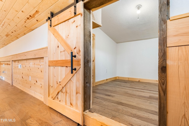 additional living space with a textured ceiling, wood walls, a barn door, light hardwood / wood-style floors, and vaulted ceiling