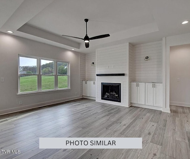 unfurnished living room with a tray ceiling, a large fireplace, ceiling fan, and light wood-type flooring