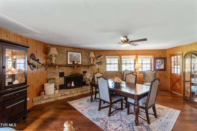 dining area with ceiling fan, dark hardwood / wood-style flooring, wood walls, and a brick fireplace