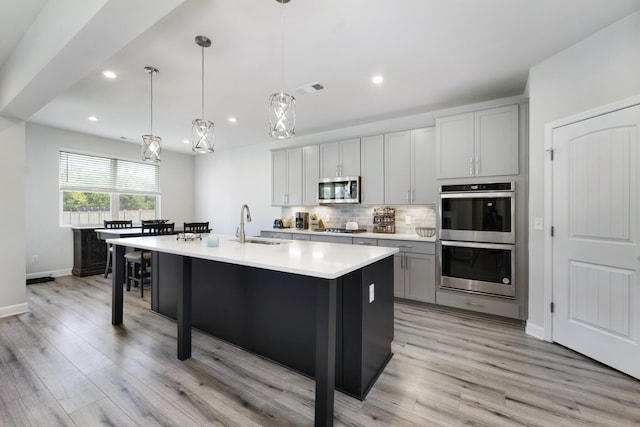 kitchen with hanging light fixtures, decorative backsplash, an island with sink, appliances with stainless steel finishes, and light hardwood / wood-style flooring