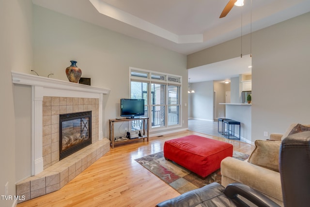 living room featuring a tray ceiling, a tiled fireplace, light hardwood / wood-style flooring, and ceiling fan