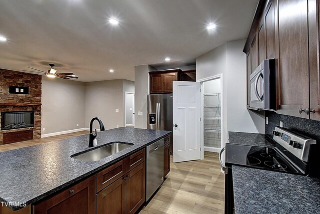 kitchen featuring stainless steel appliances, a stone fireplace, light wood-type flooring, an island with sink, and ceiling fan