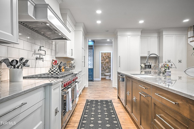 kitchen with wall chimney exhaust hood, sink, white cabinetry, light stone counters, and appliances with stainless steel finishes