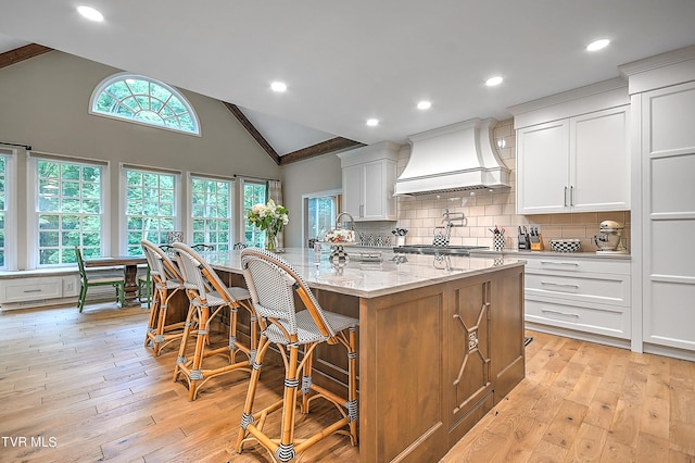 kitchen with a breakfast bar area, white cabinets, light stone countertops, custom range hood, and a spacious island