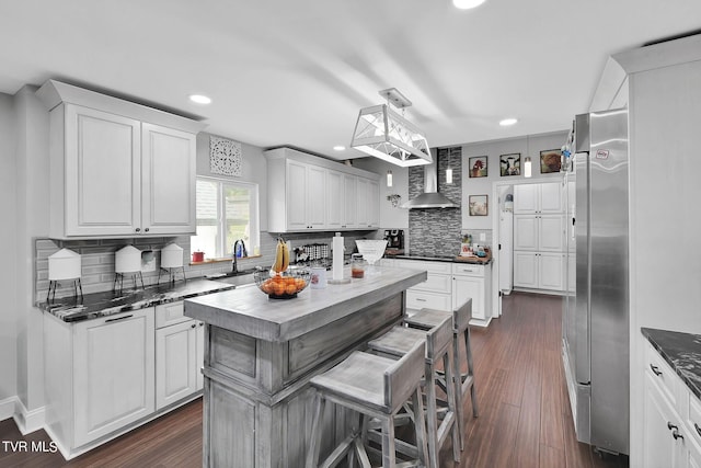 kitchen featuring white cabinets, wall chimney range hood, a center island, stainless steel built in refrigerator, and decorative light fixtures