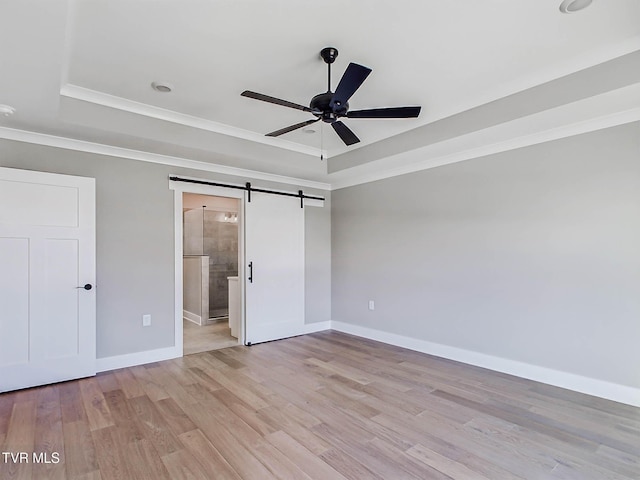 unfurnished bedroom featuring connected bathroom, ceiling fan, a barn door, a tray ceiling, and light wood-type flooring