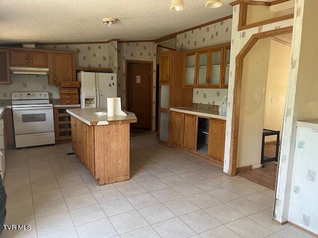 kitchen with white appliances, brown cabinetry, wallpapered walls, ornamental molding, and under cabinet range hood