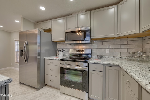 kitchen with gray cabinetry, backsplash, crown molding, appliances with stainless steel finishes, and light stone countertops