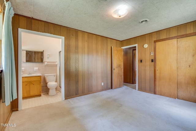 unfurnished bedroom featuring a closet, ensuite bath, sink, light colored carpet, and wood walls