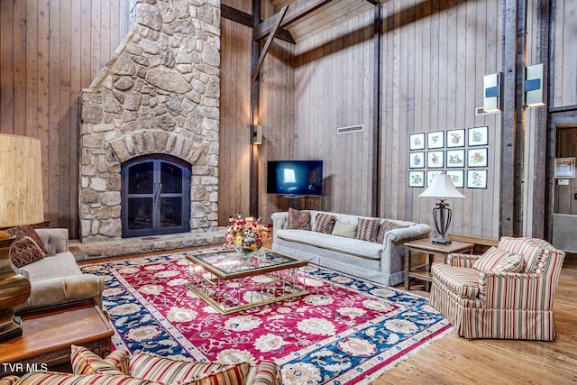 living room with a stone fireplace, wood-type flooring, a towering ceiling, wood walls, and beam ceiling