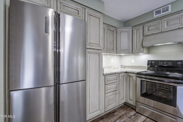 kitchen with cream cabinetry, a textured ceiling, stainless steel appliances, light stone counters, and dark wood-type flooring