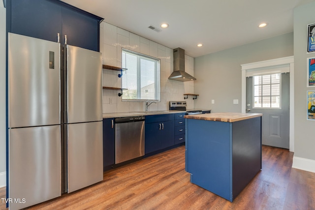 kitchen with butcher block counters, a kitchen island, wall chimney exhaust hood, appliances with stainless steel finishes, and blue cabinets