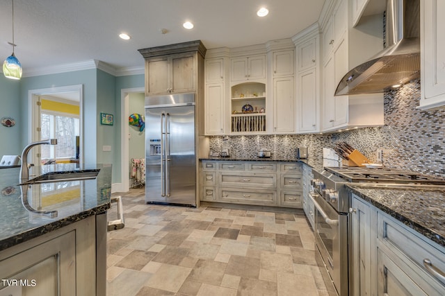 kitchen with pendant lighting, dark stone counters, sink, high end appliances, and wall chimney exhaust hood