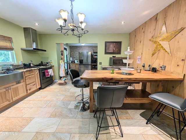 kitchen featuring pendant lighting, wooden walls, wall chimney exhaust hood, stainless steel appliances, and a notable chandelier