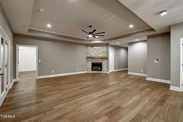 unfurnished living room featuring a stone fireplace, a raised ceiling, ceiling fan, and light wood-type flooring