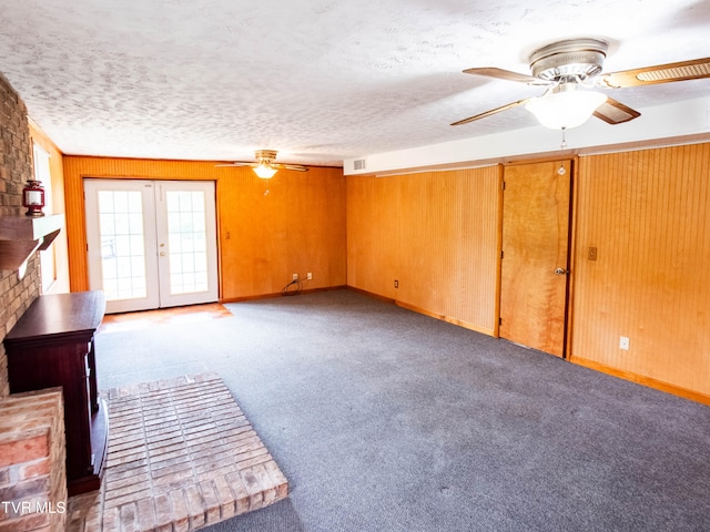 unfurnished living room with a textured ceiling, carpet flooring, wood walls, ceiling fan, and french doors