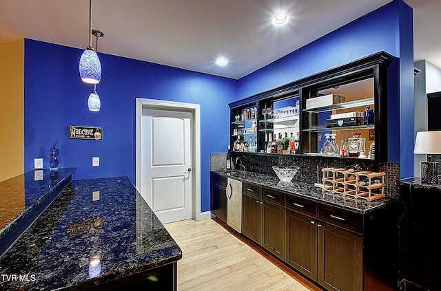 bar with sink, dark brown cabinets, decorative light fixtures, dark stone counters, and light wood-type flooring