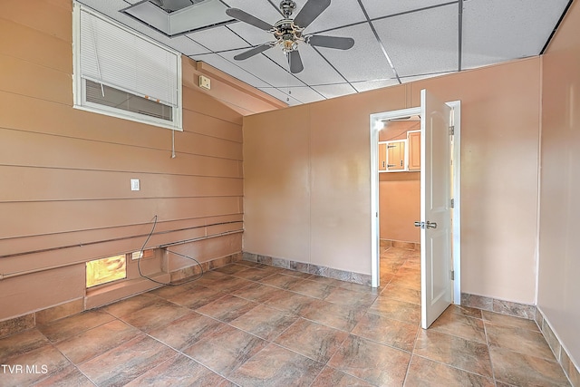 empty room with a paneled ceiling, baseboards, a ceiling fan, wood walls, and stone finish flooring