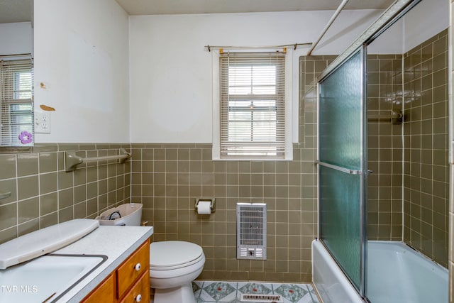 full bathroom with toilet, tile walls, heating unit, shower / bath combination with glass door, and vanity