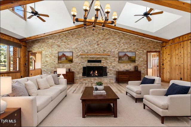 living area with beam ceiling, ceiling fan with notable chandelier, wood finished floors, a stone fireplace, and wood walls