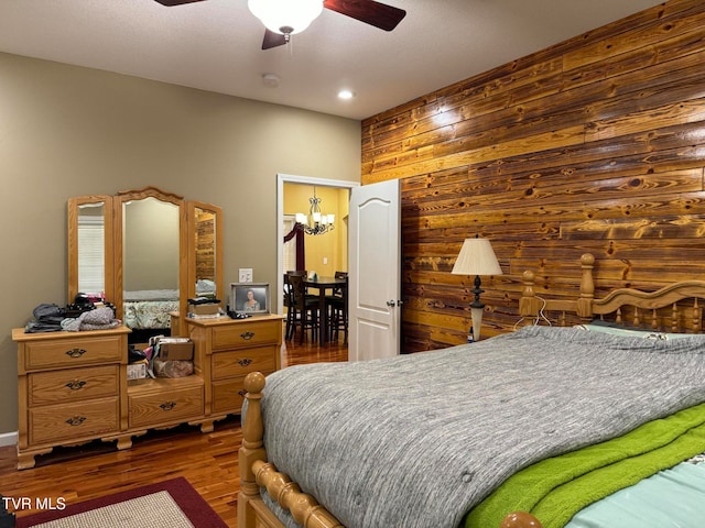 bedroom featuring ceiling fan with notable chandelier, dark wood-type flooring, and wooden walls