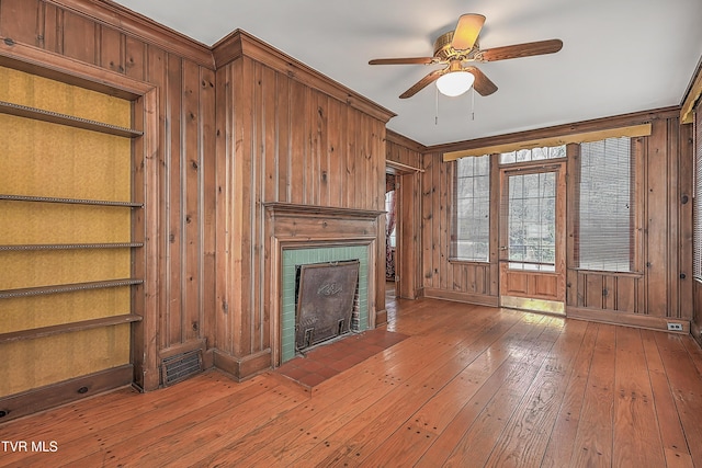 unfurnished living room with ceiling fan, crown molding, wooden walls, wood-type flooring, and a fireplace